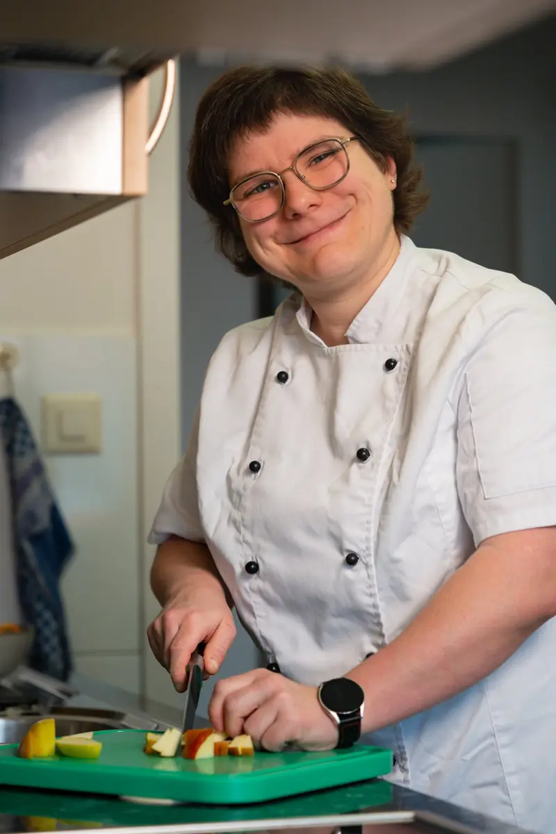 Anita Arpagaus in weisser Kochjacke schneidet in ihrer Küche Apfelspalten auf einem grünen Brett und lächelt in die Kamera