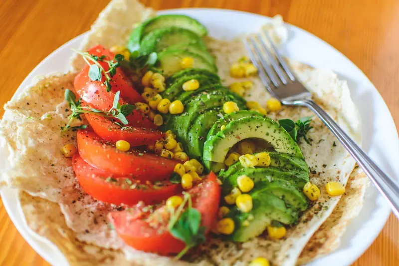 Weisser Teller mit einer Tortilla, belegt mit Tomatenscheiben, Avocado, Mais und frischen Kräutern