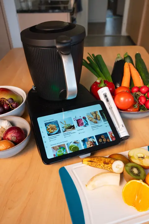 Thermomix und Tablet auf einem Holztisch, davor ein Schneidebrett mit halbierten Früchten