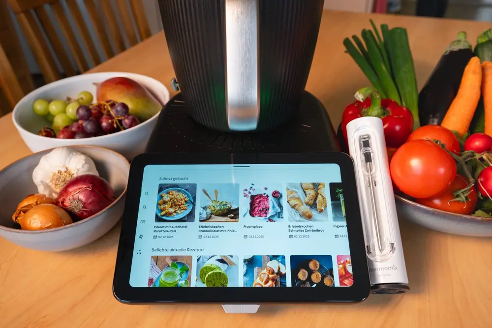 Thermomix auf einem Holztisch, davor ein Tablet mit Rezepten, daneben Schalen mit Früchten, Zwiebeln und frischem Gemüse